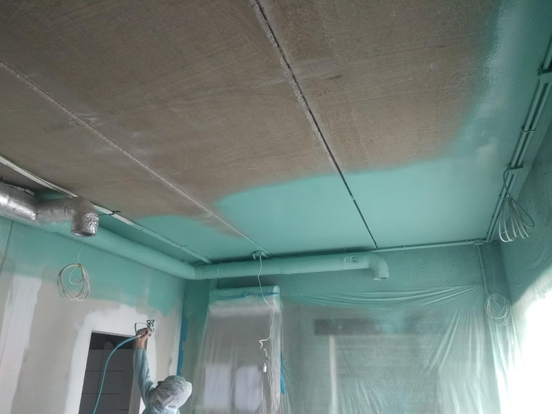 Prace malarskie w pomieszczeniu: malowanie sufitu natryskowo na kolor turkusowy, widoczny fragment sufitu przed malowaniem w kolorze szarym, rury wentylacyjne, zabezpieczone okna folią malarską.