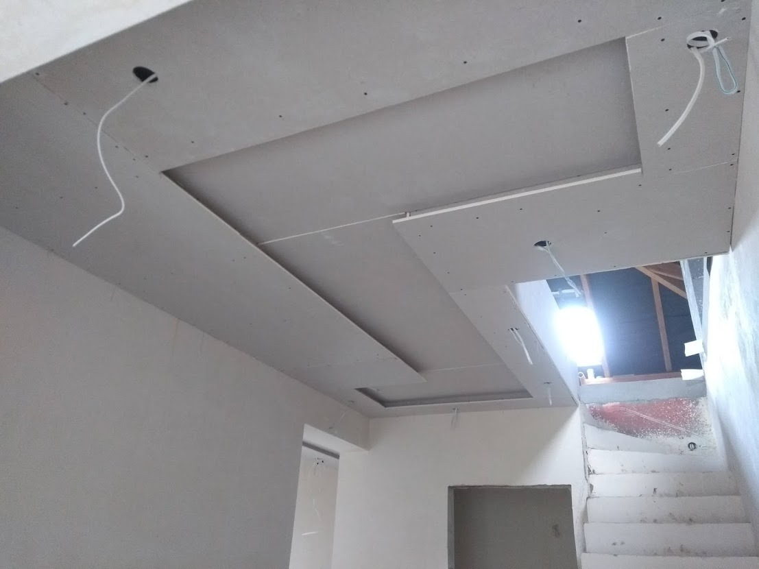 Sufit z płyt kartonowo-gipsowych w trakcie montażu, widoczne przewody elektryczne i otwory pod oświetlenie, surowe wykończenie wnętrza przy schodach.