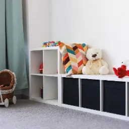 Umeblowane wnętrze pokoju dziecięcego z białymi regałami wypełnionymi zabawkami, poduszkami i wózkiem dla lalek z wikliny.