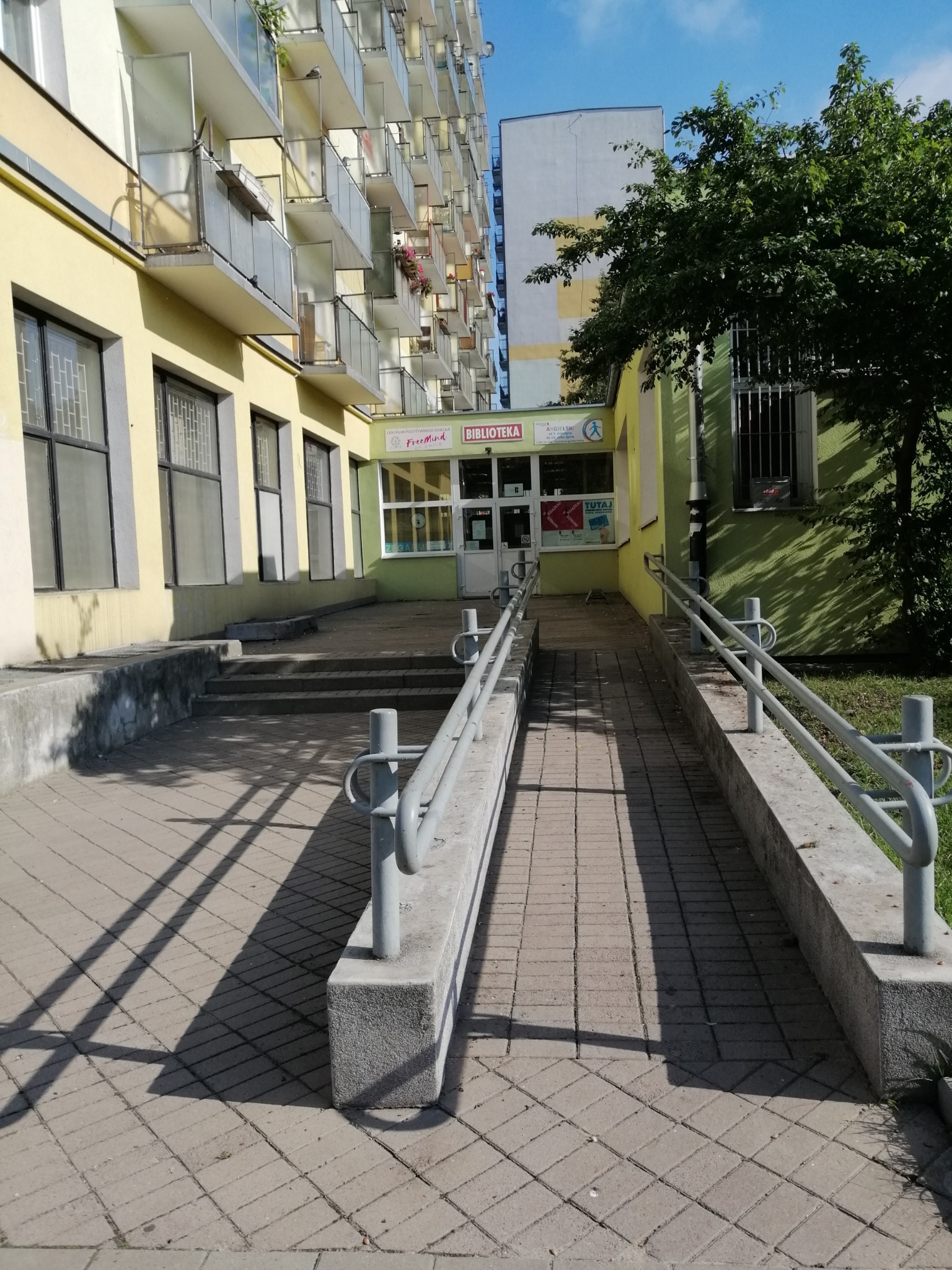 Wejście do budynku z rampą dla wózków, nad wejściem szyld z napisem BIBLIOTEKA oraz logo Centrum Pozytywnego Dziecka FreeMIND, w tle budynek mieszkalny z balkonami.