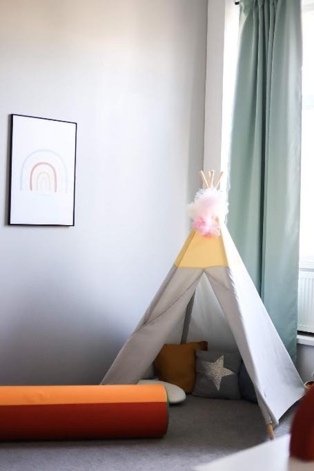 Pokój dziecięcy w Łodzi z tipi w odcieniach szarości i żółci, ozdobionym różowym pomponem, obok pomarańczowy wałek do ćwiczeń i plakat z tęczą.