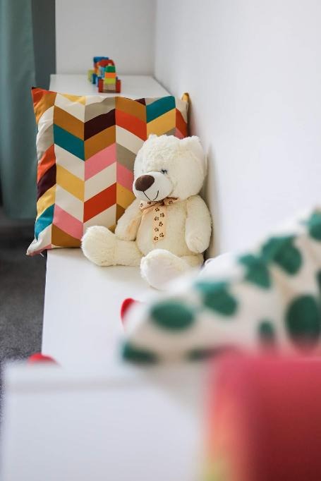 Jasny pokój dziecięcy z pluszowym misiem siedzącym obok poduszki w geometryczne wzory, na tle klocki i częściowo widoczna inna poduszka w grochy.