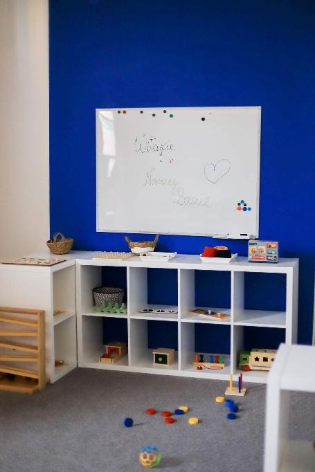 Sala żłobkowa z białymi regałami wypełnionymi zabawkami i pomocami edukacyjnymi, na ścianie tablica z dziecięcymi rysunkami i napisami, na podłodze rozrzucone kolorowe klocki i piłeczka.