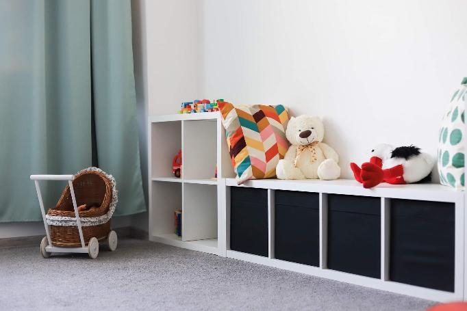 Umeblowane wnętrze pokoju dziecięcego z białymi regałami wypełnionymi zabawkami, poduszkami i wózkiem dla lalek z wikliny.