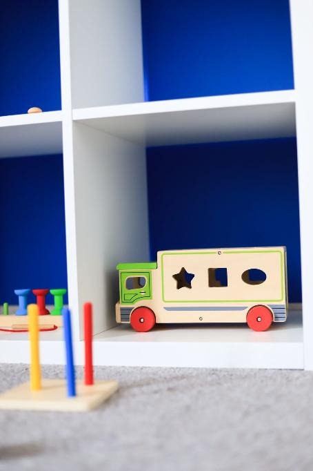 Drewniana ciężarówka-sorter kształtów i wieża edukacyjna z kolorowymi krążkami w białym regale na tle niebieskiej ściany.