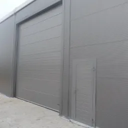 Szara brama przemysłowa segmentowa z bocznymi drzwiami wejściowymi, widoczne poziome przetłoczenia na panelach, szara kostka brukowa przed budynkiem.