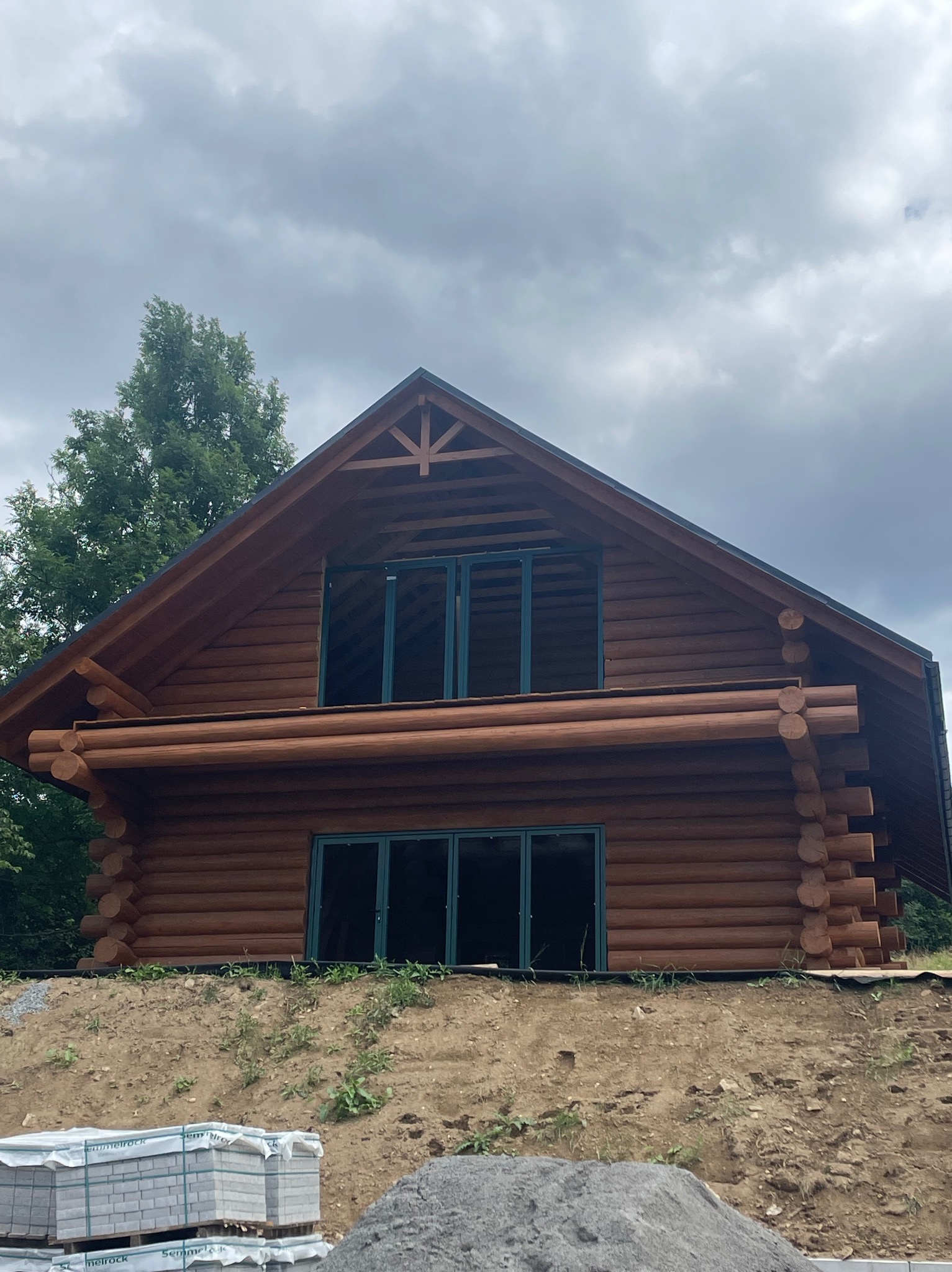 Dom z bali w stylu góralskim z ciemnozielonymi oknami, położony na wzniesieniu, widoczne materiały budowlane.