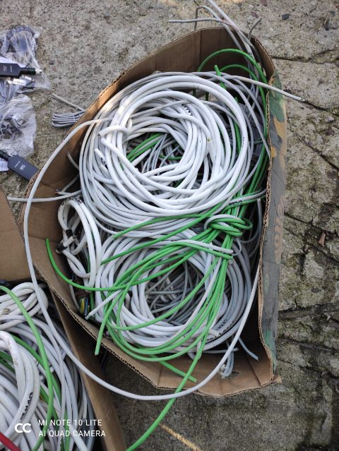 Karton z kablami telekomunikacyjnymi: białe i zielone przewody zwinięte w kręgi, leżące na szarym betonie obok czarnych adapterów sieciowych.