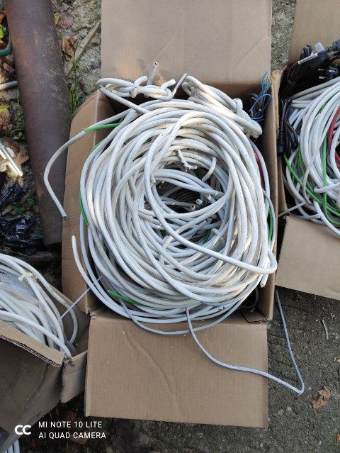 Kartonowe pudło wypełnione zwiniętymi kablami elektrycznymi w białej izolacji, widoczne końcówki kabli i inne kable w tle.