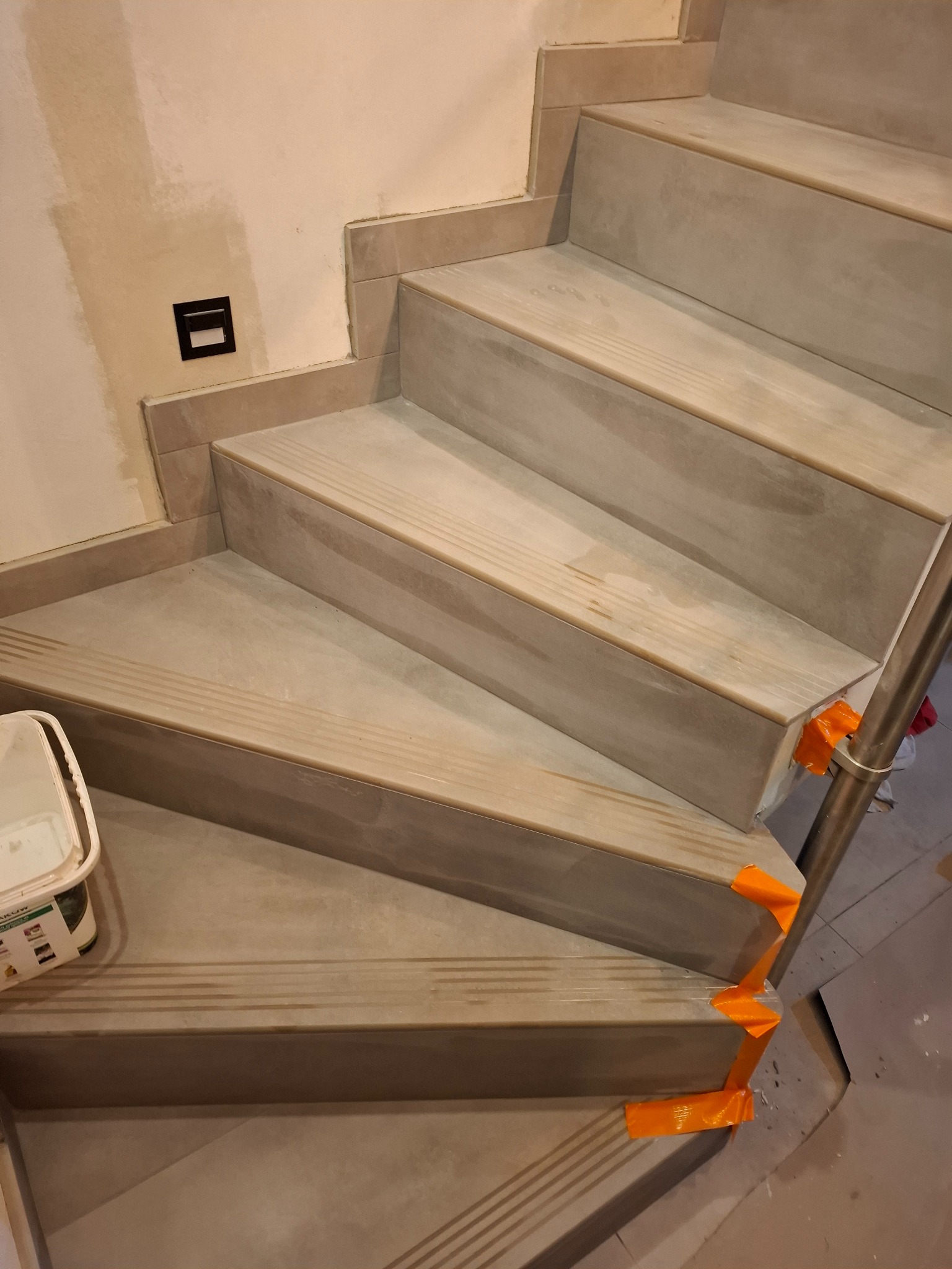 Wykończone schody z szarych płytek z antypoślizgowymi rowkami, zabezpieczone pomarańczową taśmą malarską na krawędziach. Widoczna puszka z farbą i fragment ściany z gniazdkiem.