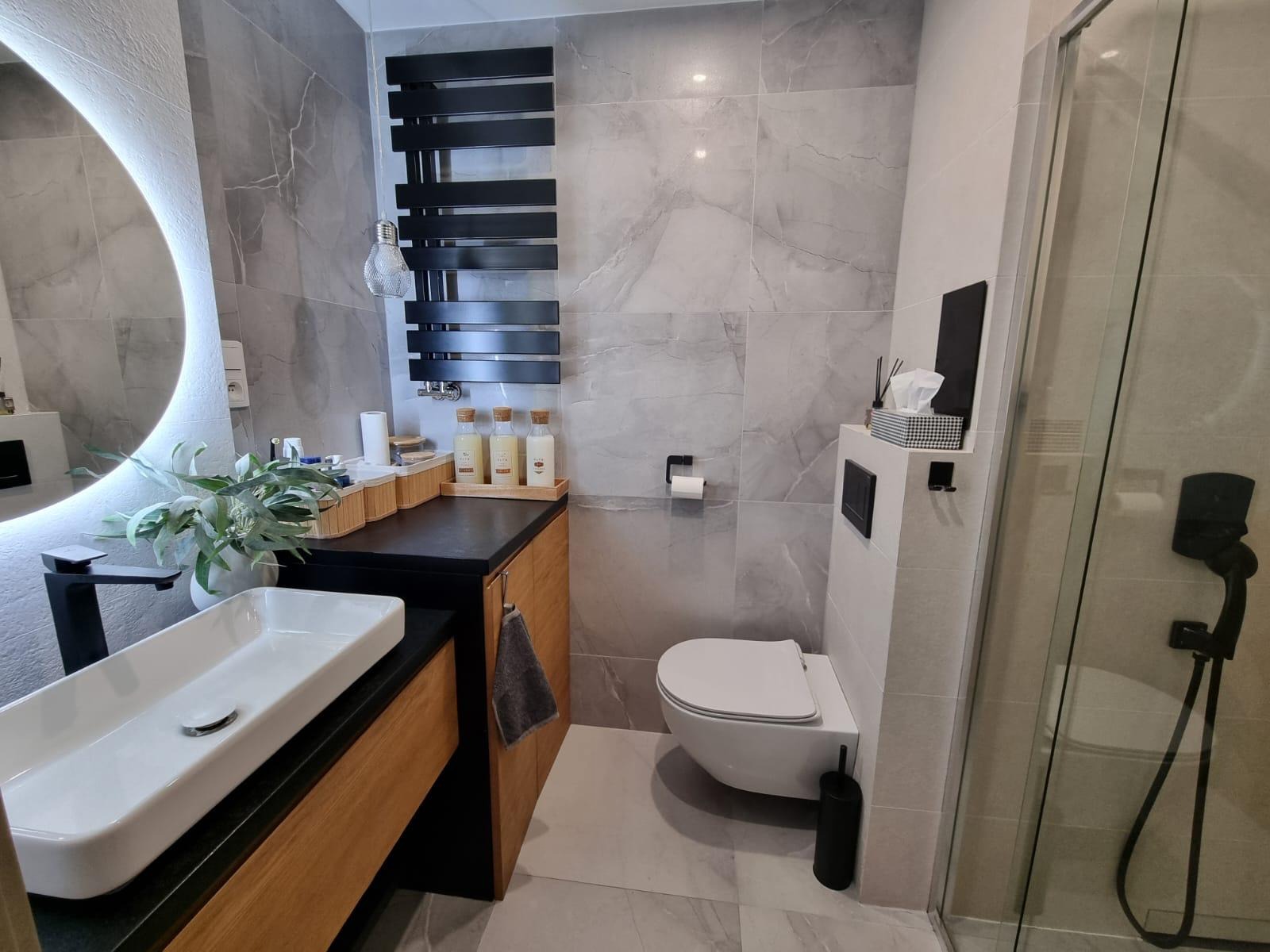 Nowoczesna łazienka z marmurowymi płytkami, podświetlanym lustrem, drewnianą szafką pod umywalką i czarnym grzejnikiem drabinkowym. Minimalistyczny design.