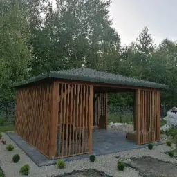 Drewniana konstrukcja z pionowymi listwami i płaskim dachem, otoczona zielenią i jasnym żwirem, z betonową posadzką wewnątrz.
