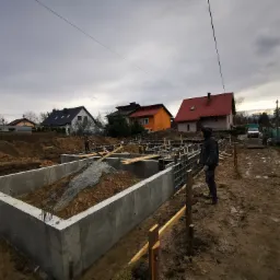 Wykopane fundamenty domu jednorodzinnego, zalane betonem, z widocznym zbrojeniem i szalunkami, częściowo zasypane ziemią i żwirem, na tle zabudowy jednorodzinnej pod pochmurnym niebem. Pracownik...
