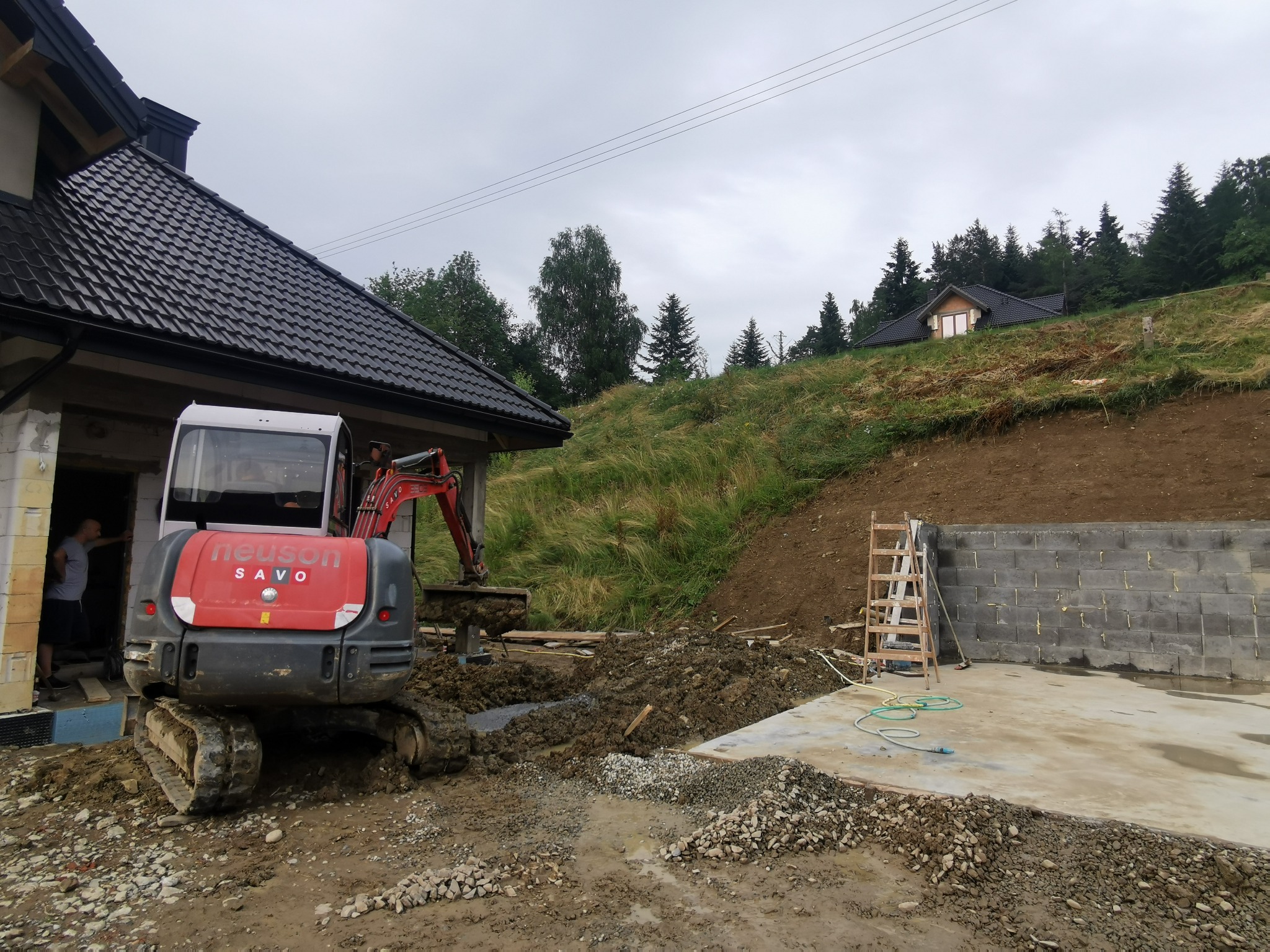 Czerwono-szara mini koparka Neuson SAVO na błotnistym terenie budowy, w tle wzniesiona ściana z betonowych bloczków, drewniana drabina i dom na wzgórzu pod pochmurnym niebem.