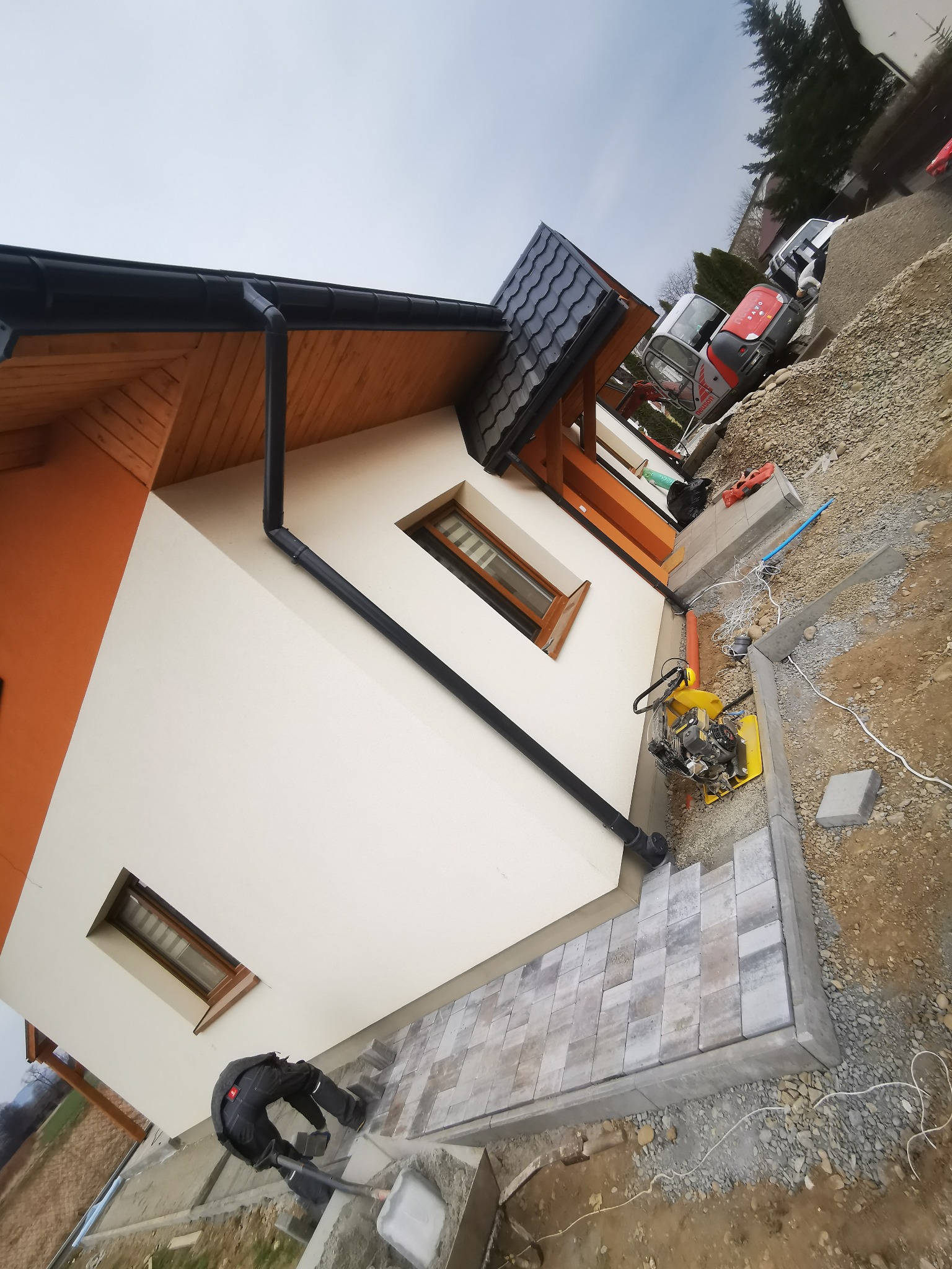 Układanie kostki brukowej wokół nowo wybudowanego domu z pomarańczowo-białą elewacją, widoczny zagęszczarka wibracyjna i mini koparka w tle.