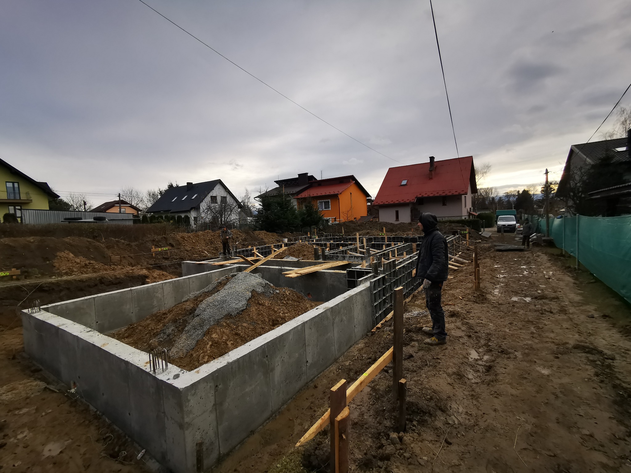 Wykopane fundamenty domu jednorodzinnego, zalane betonem, z widocznym zbrojeniem i szalunkami, częściowo zasypane ziemią i żwirem, na tle zabudowy jednorodzinnej pod pochmurnym niebem. Pracownik...