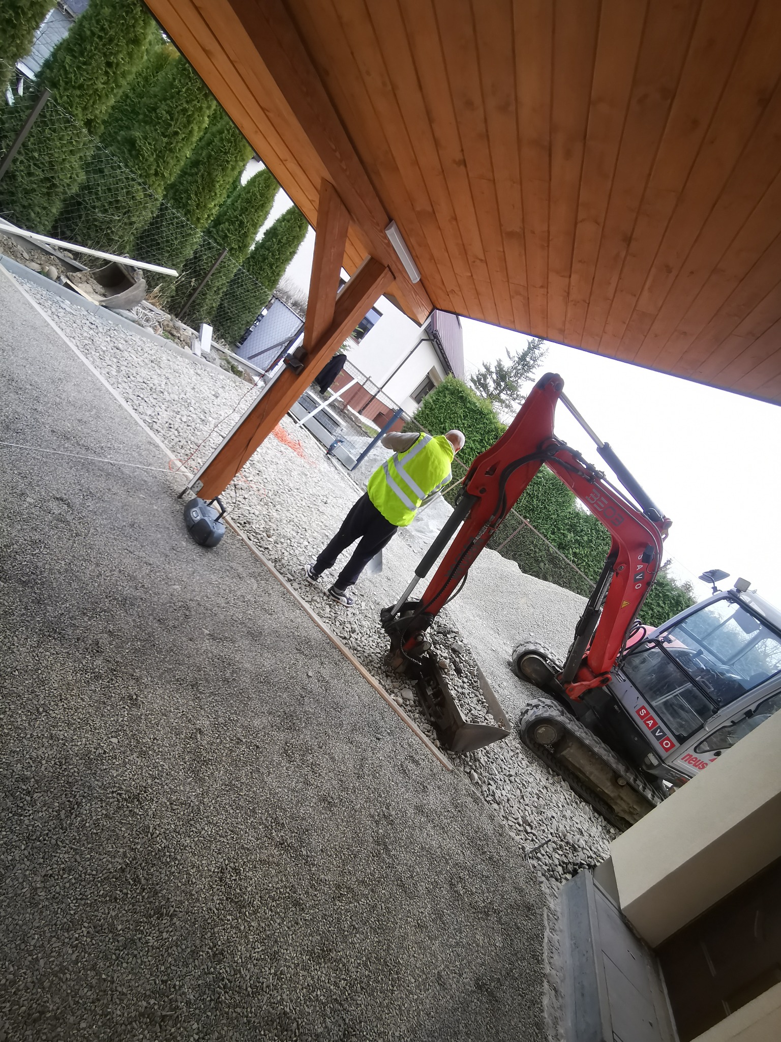 Czerwony mini-koparka SAVO na żwirowym podłożu, pracownik w żółtej kamizelce obok, widok spod drewnianej konstrukcji z zewnątrz budynku.