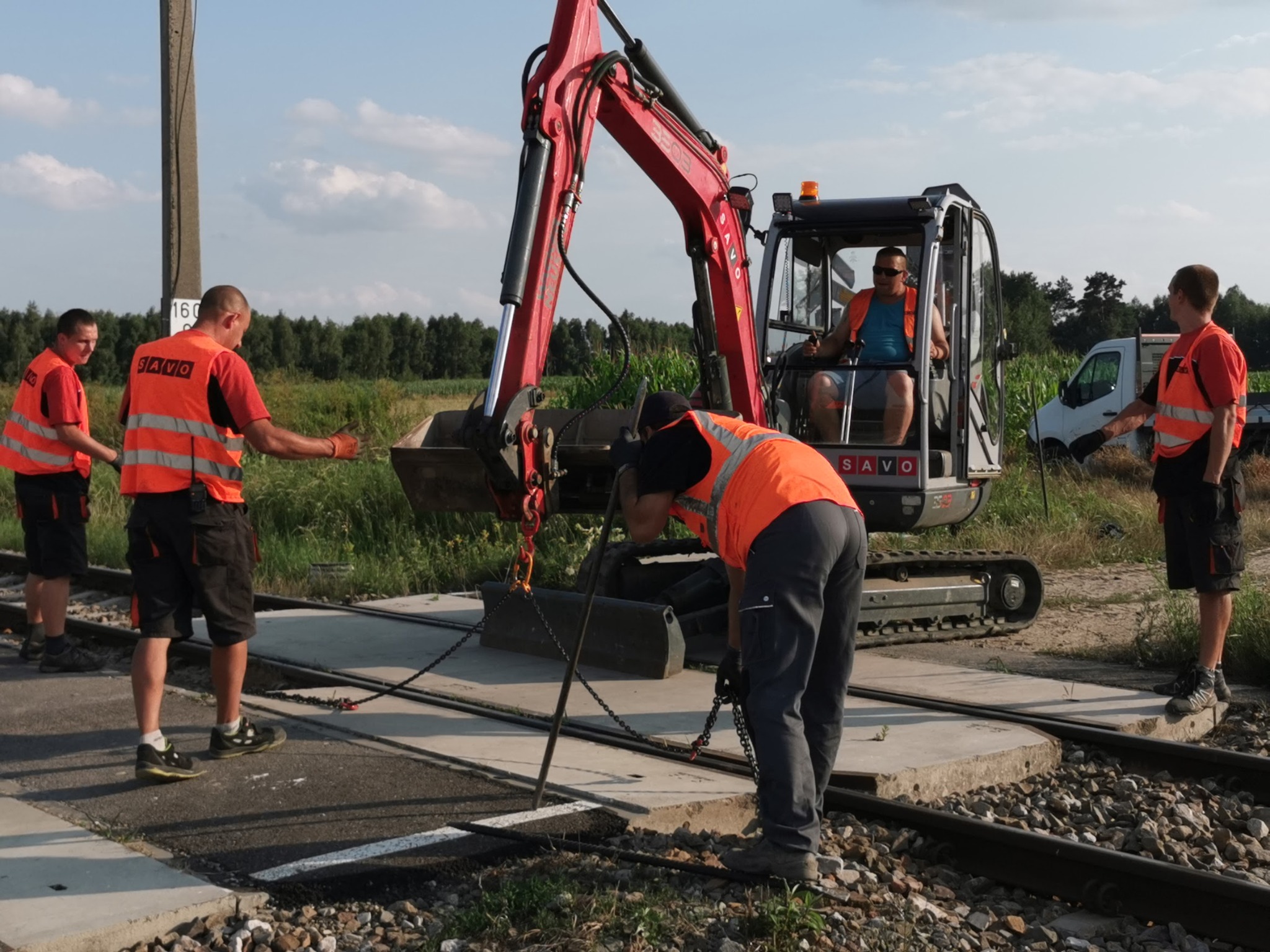 Pracownicy w kamizelkach odblaskowych przy czerwonej mini koparce, podnoszącej betonowy element na torach kolejowych, w tle zieleń i biały samochód dostawczy.