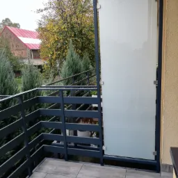 Szklana balustrada balkonowa z metalową ramą, widok z zewnątrz, Połajewo. W tle budynek z czerwoną dachówką i jesienne drzewa.