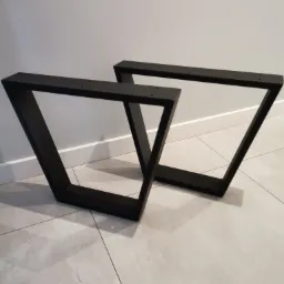 Dwie czarne, metalowe nogi do stołu o trapezoidalnym kształcie, leżące na szarych płytkach podłogowych, z widocznymi otworami montażowymi.