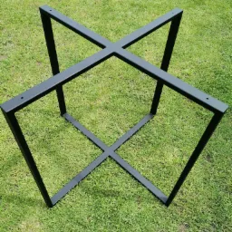 Metalowa, spawana podstawa stołu w kształcie krzyża, pomalowana na czarno, leżąca na zielonej trawie w słoneczny dzień.