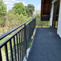 Czarna, metalowa barierka balkonowa z widokiem na zieleń i błękitne niebo, zamontowana na balkonie wykończonym czarną papą, widoczny fragment budynku z drewnianą stolarką okienną.