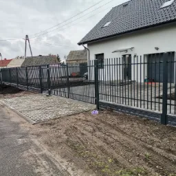 Nowoczesna, metalowa konstrukcja ogrodzenia posesji z bramą przesuwną, widoczna na tle białego domu z szarą dachówką i kostki brukowej. Teren wokół ogrodzenia częściowo uporządkowany, widoczne...