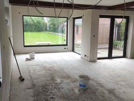 Wnętrze w trakcie szpachlowania: widoczne ślady szpachli na podłodze, ściany przygotowane do dalszych prac, puszki z materiałem i narzędzia w narożniku, duże okno z widokiem na ogród.