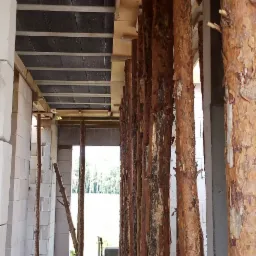 Wnętrze surowego domu w trakcie budowy z widocznymi ścianami z białych bloczk&oacute;w, betonowym stropem podpartym drewnianymi stemplami z korą i prowizorycznym oknem z widokiem na las.