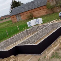 Wykopany fundament z izolacją bitumiczną, bloczki betonowe, drewniane szalunki, widok na budynek gospodarczy z cegły i pole w tle.