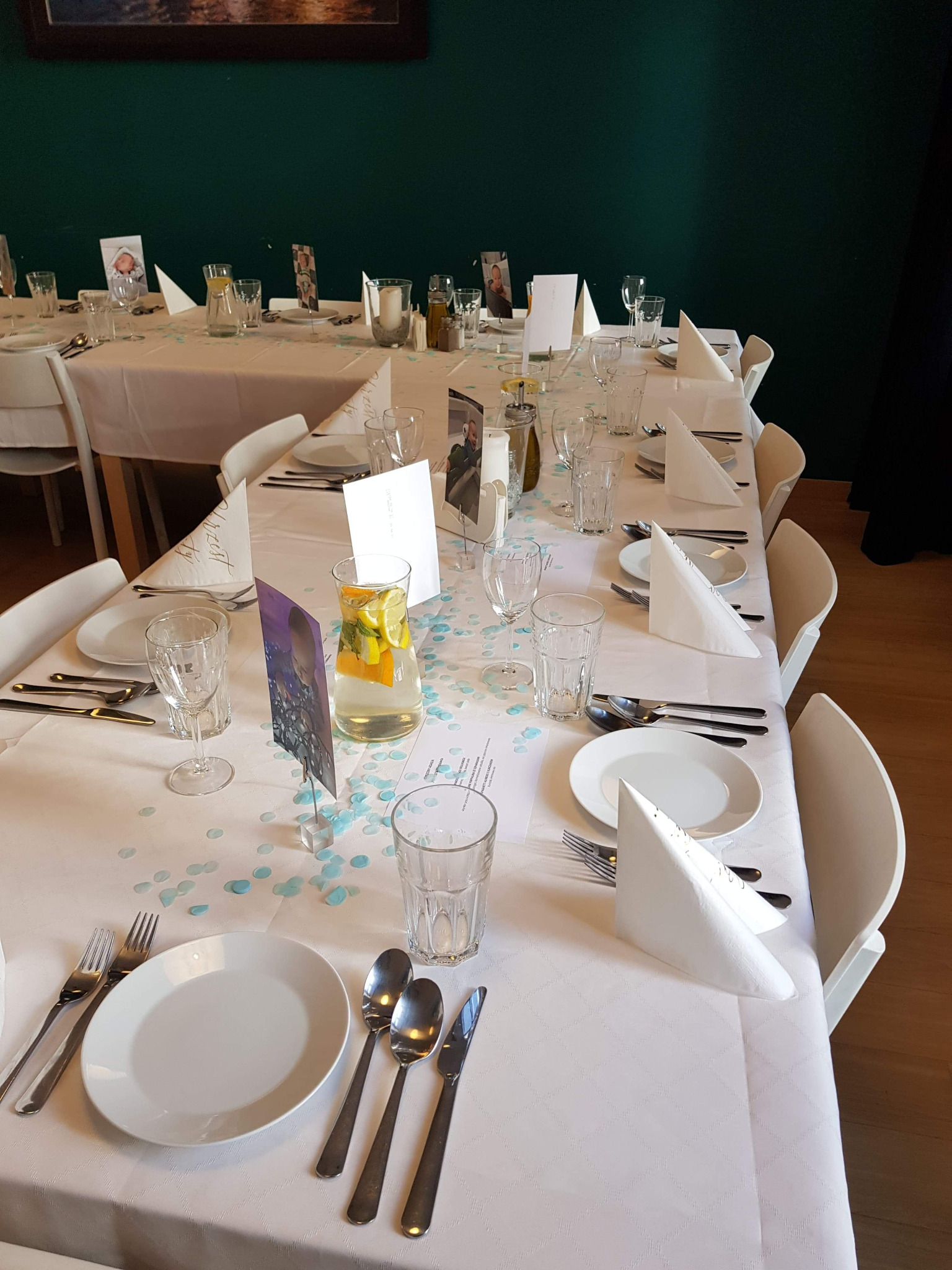 Elegancko nakryty stół z białym obrusem w delikatny wzór, białą zastawą, szklankami, sztućcami, serwetkami ułożonymi w trójkąty i dzbankiem z wodą i plastrami cytryny i pomarańczy, udekorowany...