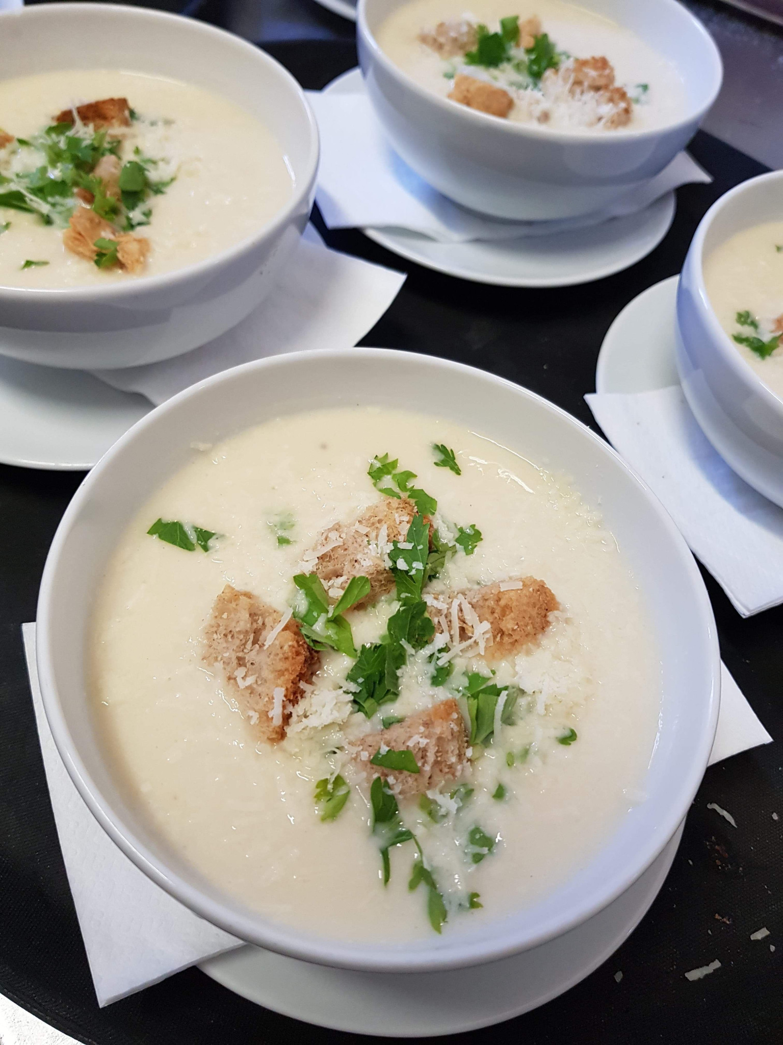 Kremowa zupa w białej misce, udekorowana grzankami, posiekaną zieloną pietruszką i startym serem, na czarnym blacie obok innych misek z zupą.