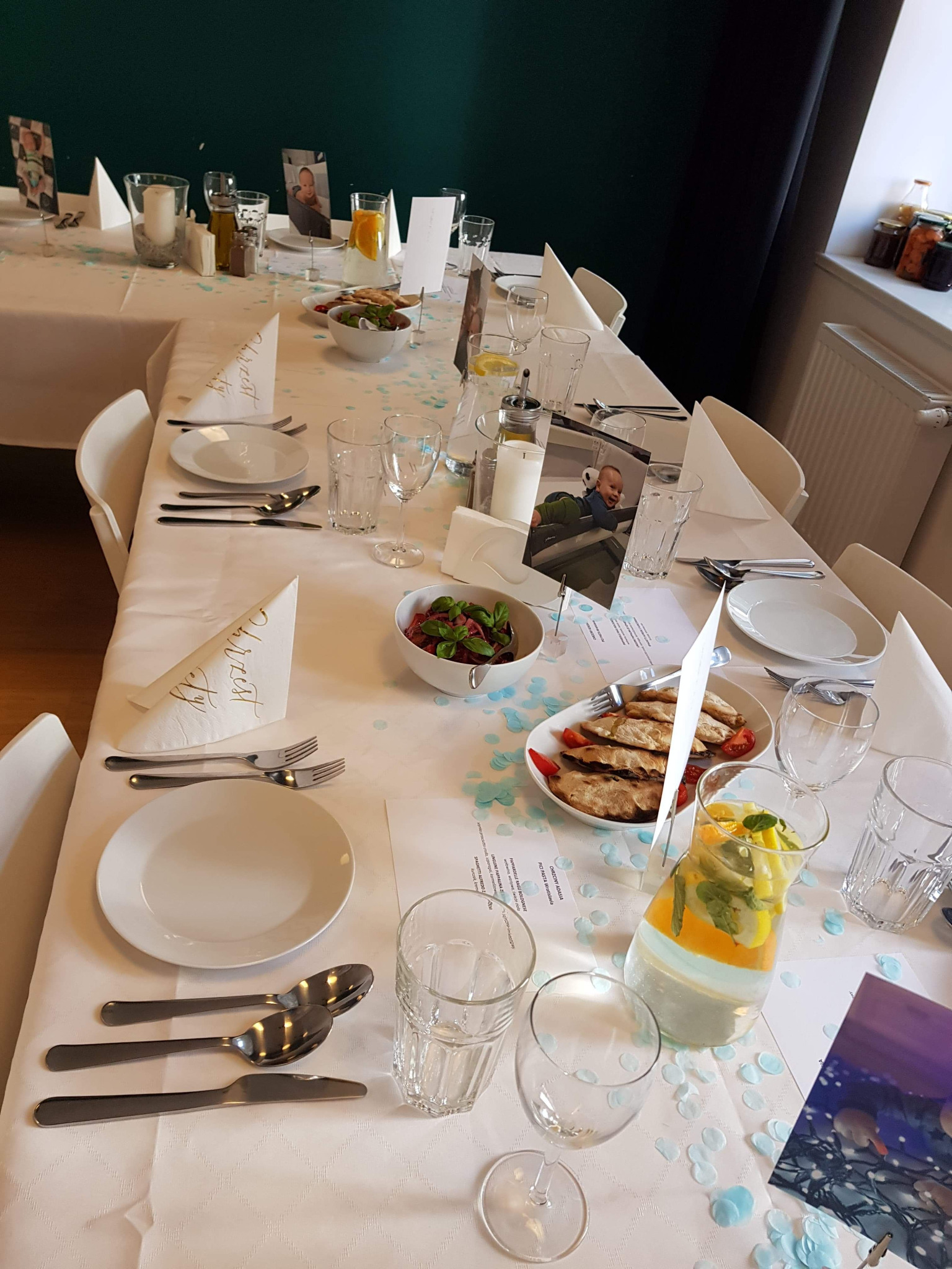 Uroczysty stół z białym obrusem i dekoracjami w odcieniach błękitu, nakryty dla gości z talerzami, sztućcami, szklankami, dzbankiem z wodą z cytryną i miętą, sałatkami i pierogami, oraz zdjęciami...