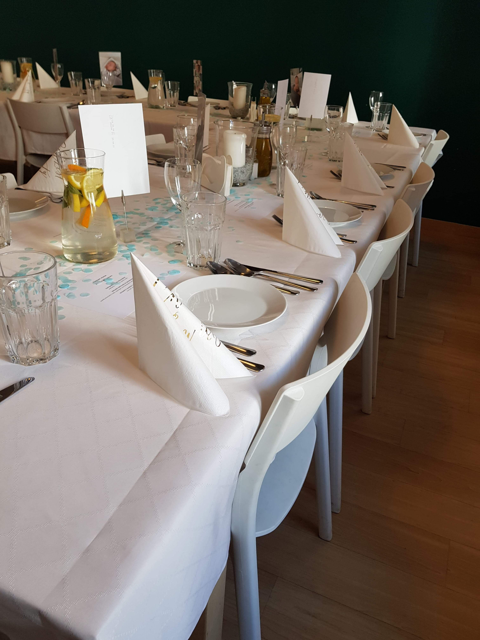 Uroczysty stół z białym obrusem w delikatny wzór, nakryty dla gości: białe talerze, sztućce, szklanki, woda z cytryną i pomarańczą w szklanym dzbanku, białe serwetki złożone w trójkąt, menu...
