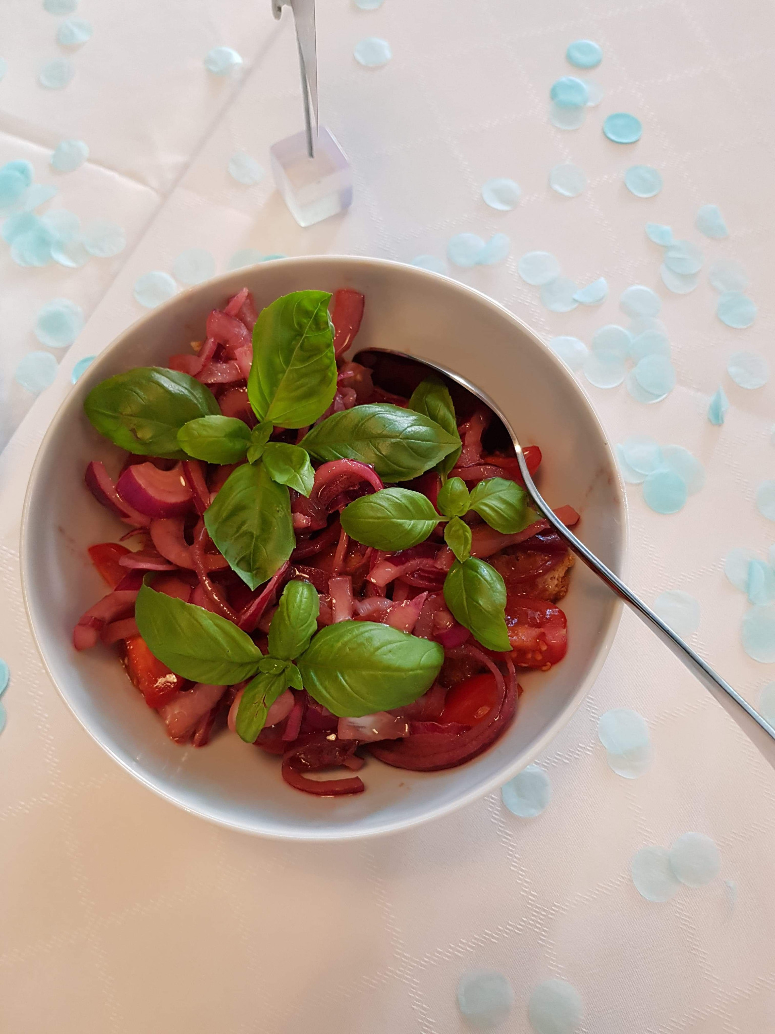 Sałatka ze świeżych pomidorów, czerwonej cebuli i bazylii w białej misce, z łyżką, na białym obrusie z dekoracyjnymi, błękitnymi konfetti.