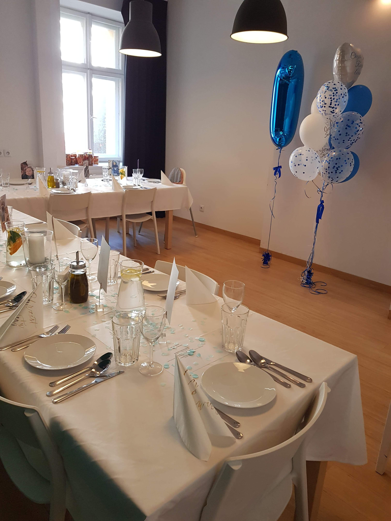 Uroczysta aranżacja stołu z białym obrusem, zastawą, szklankami, konfetti i dekoracyjnymi balonami w odcieniach bieli i błękitu, przygotowana w jasnym wnętrzu z oknem.