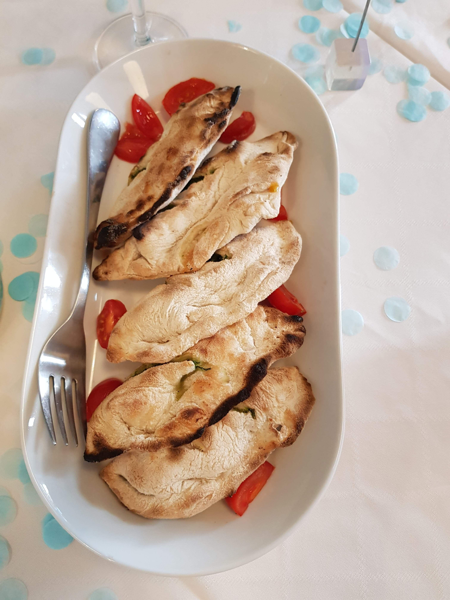 Podłużny biały talerz z pięcioma kawałkami podpłomyków z przypalonymi brzegami, udekorowanymi połówkami pomidorków koktajlowych, obok leży srebrny widelec, w tle biały obrus w błękitne kropki...