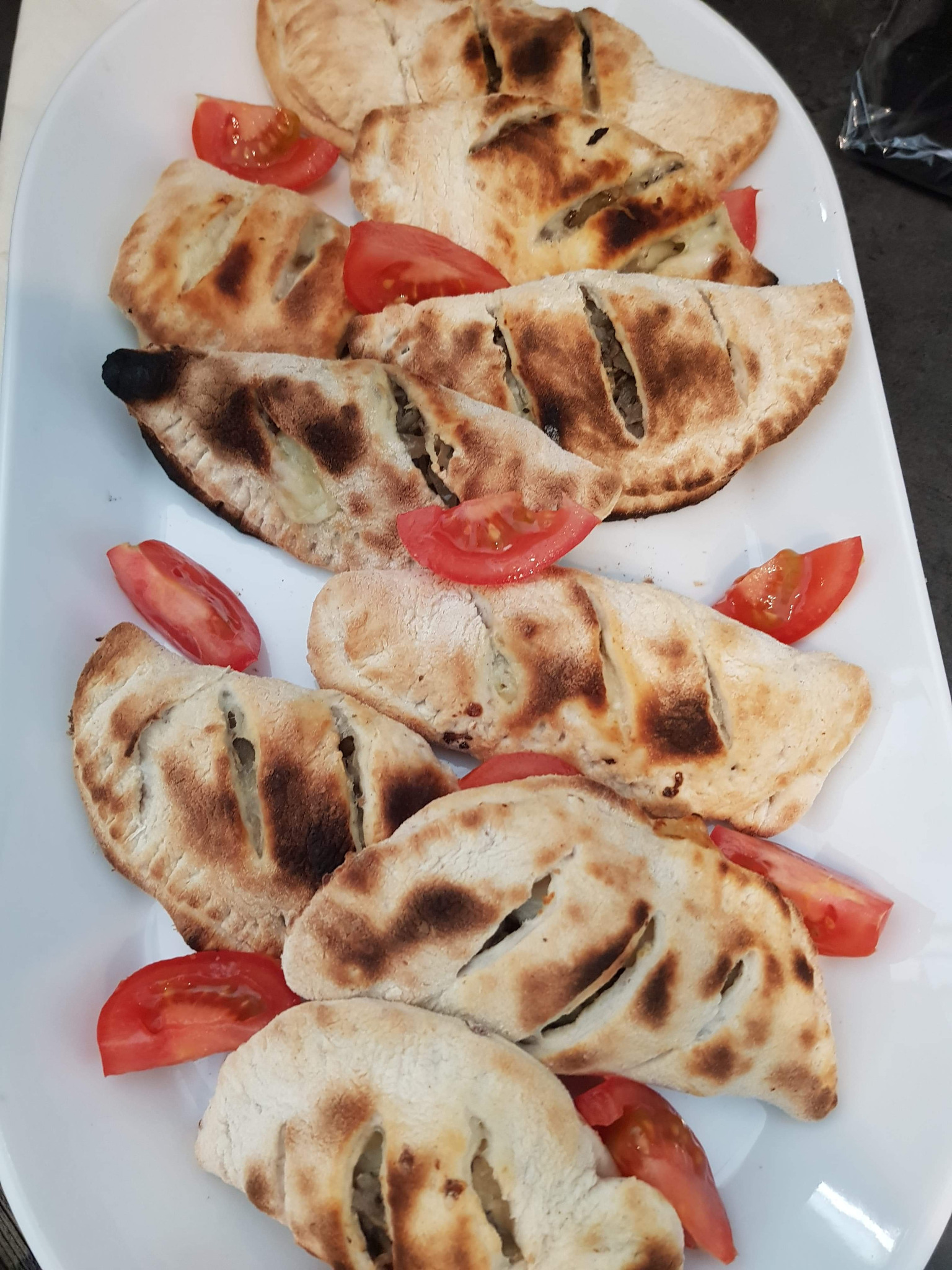 Półksiężyce z ciasta z farszem, przypominające pierogi calzone, ułożone na białym, owalnym talerzu i udekorowane ćwiartkami pomidora; jeden z nich lekko przypalony.