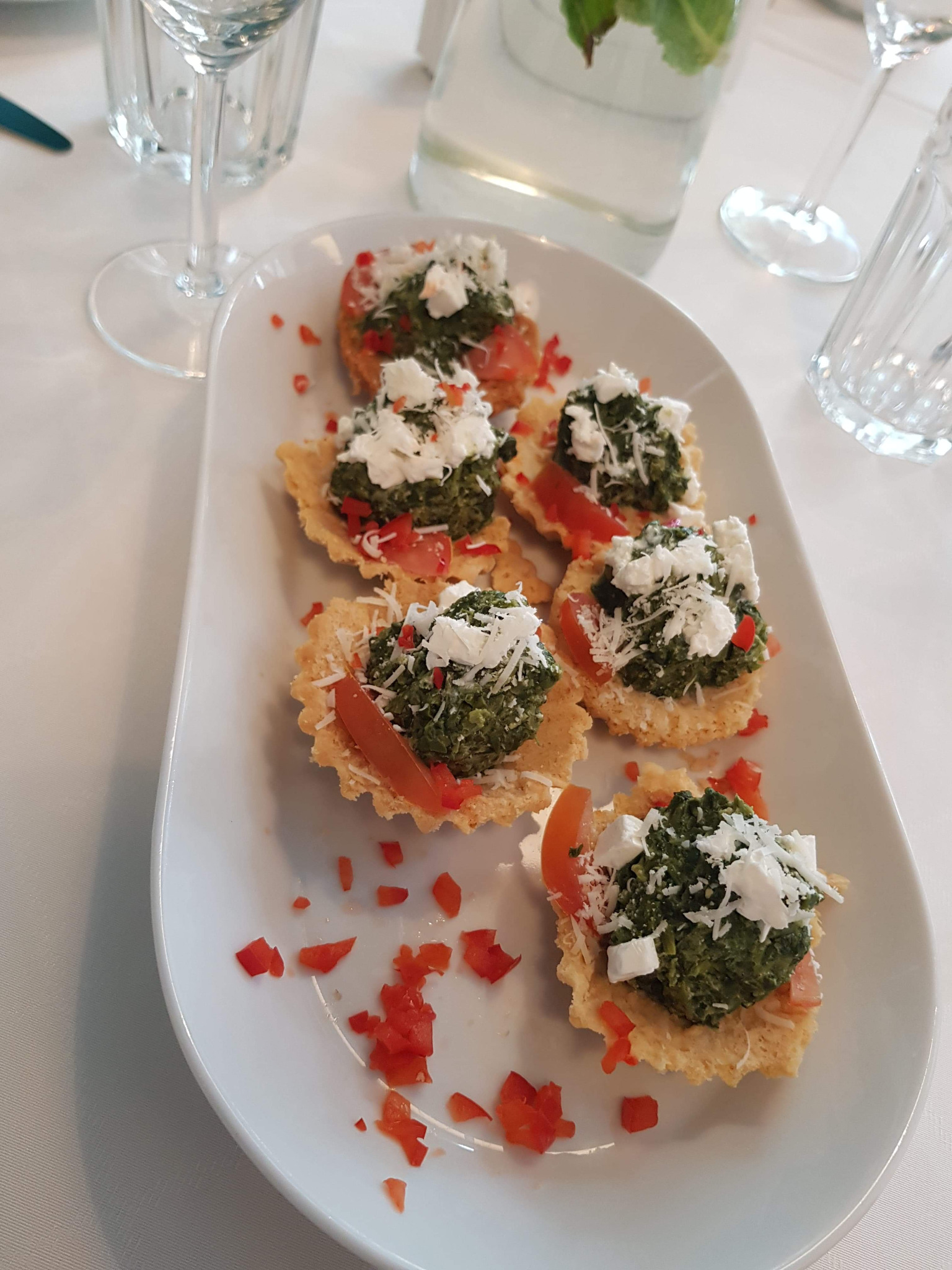 Elegancko podane tartaletki z pesto, pomidorami i serem feta na białym talerzu, widoczne kieliszki i woda z miętą w tle, jasne oświetlenie.