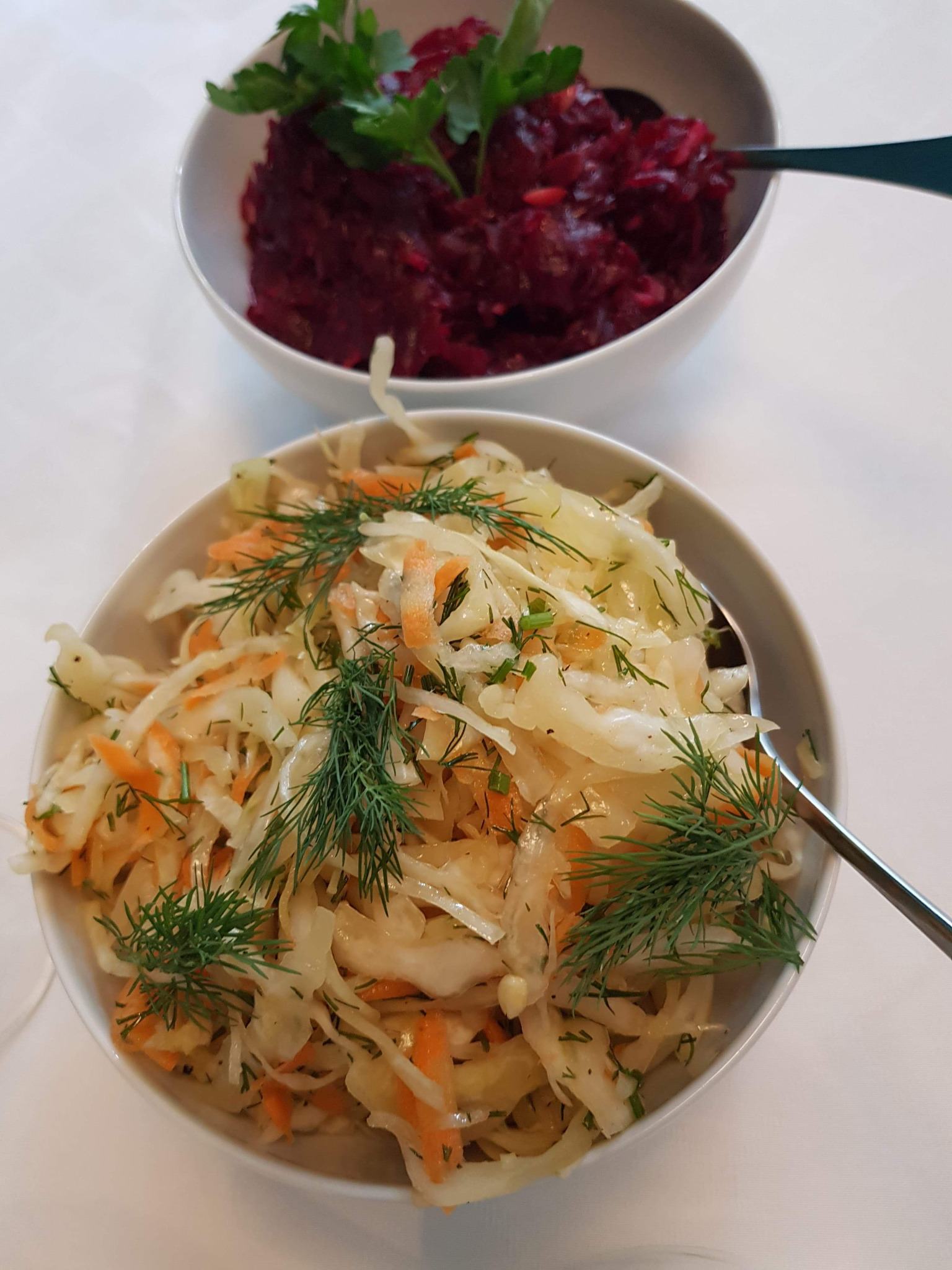 Dwie białe miski z sałatkami: surówka z kapusty z marchewką i koperkiem na pierwszym planie, sałatka z buraków z natką pietruszki w tle, obie z łyżkami.