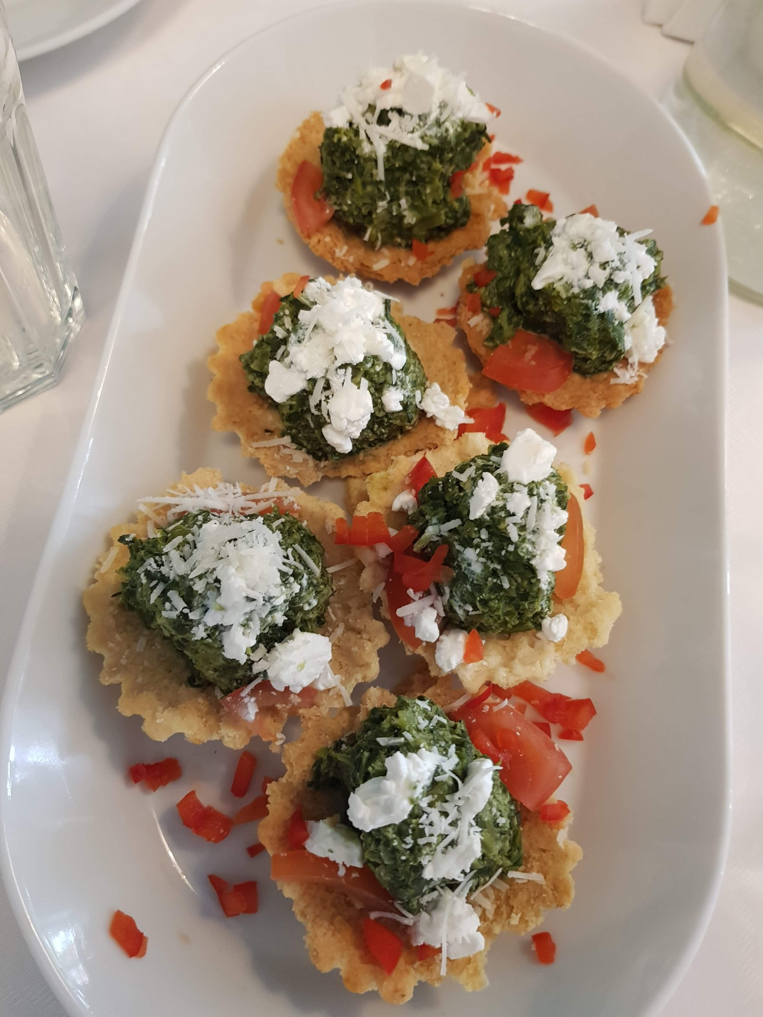 Apetizing tartlets with spinach pesto, tomato slices, and crumbled feta cheese, arranged on a white rectangular plate, next to a glass of water, showcasing a catering service.