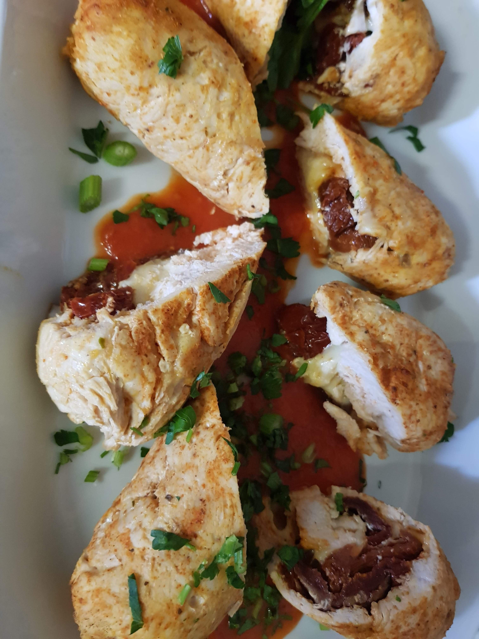 Filet z kurczaka faszerowany suszonymi pomidorami i serem, polany sosem pomidorowym i posypany zieloną pietruszką, ułożony na białym talerzu.