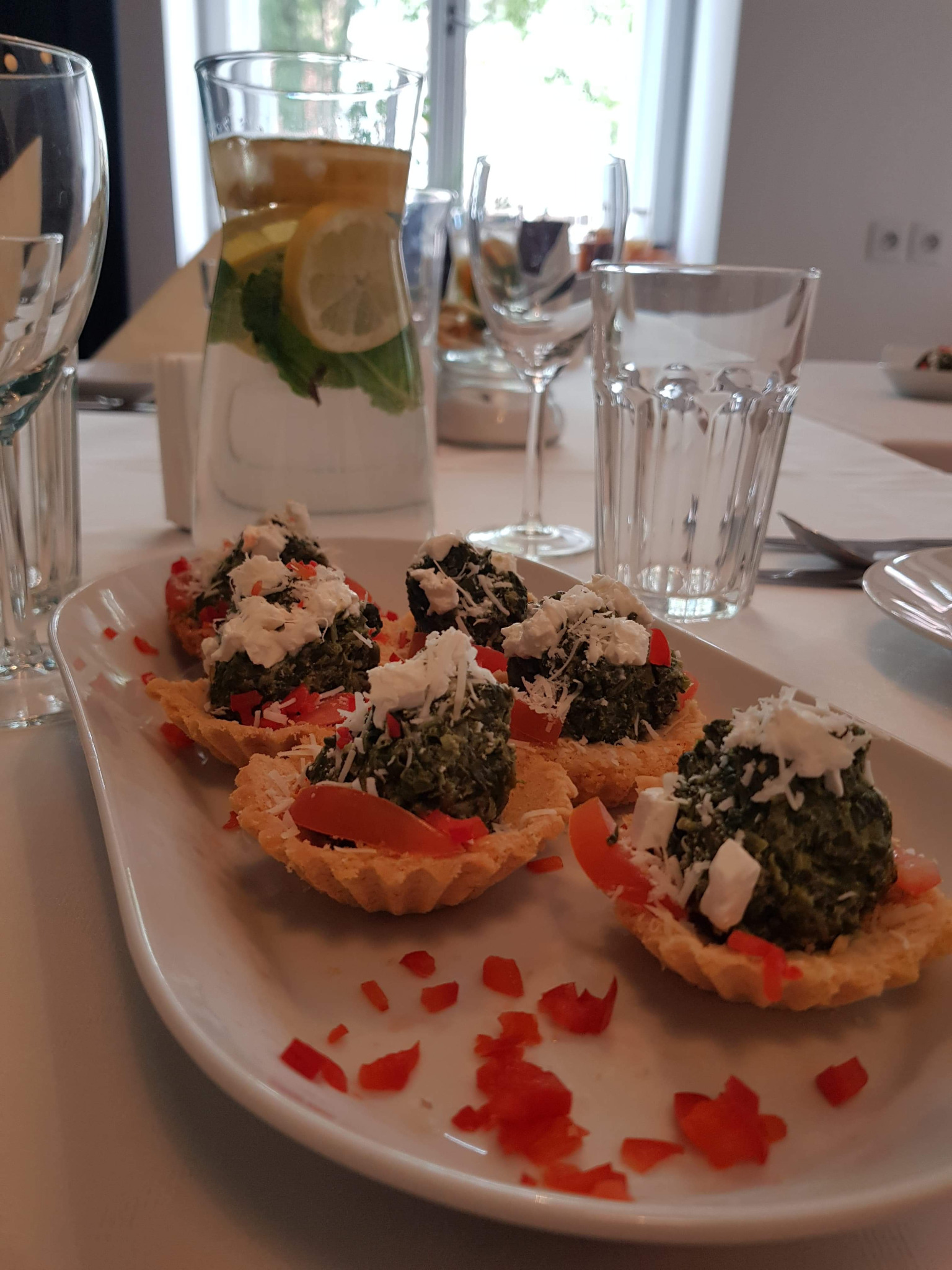 Elegancko podane tartaletki z zielonym farszem, pomidorami i serem feta na białym talerzu, w tle dzbanek z wodą, cytryną i miętą oraz nakryty stół.