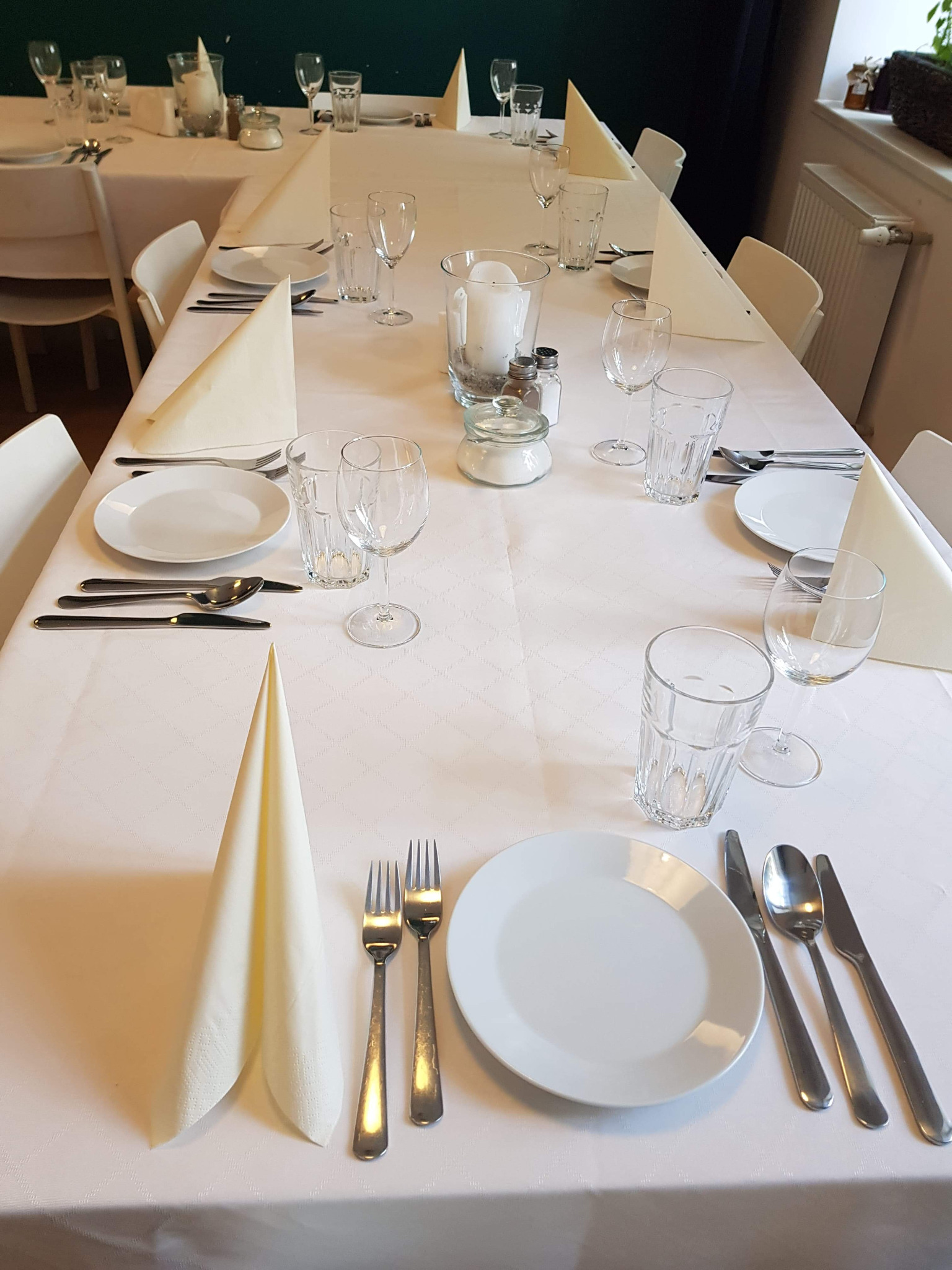 Elegancko nakryty stół z białym obrusem, talerzami, sztućcami, szklankami i serwetkami ułożonymi w stożki, przygotowany na uroczystość.