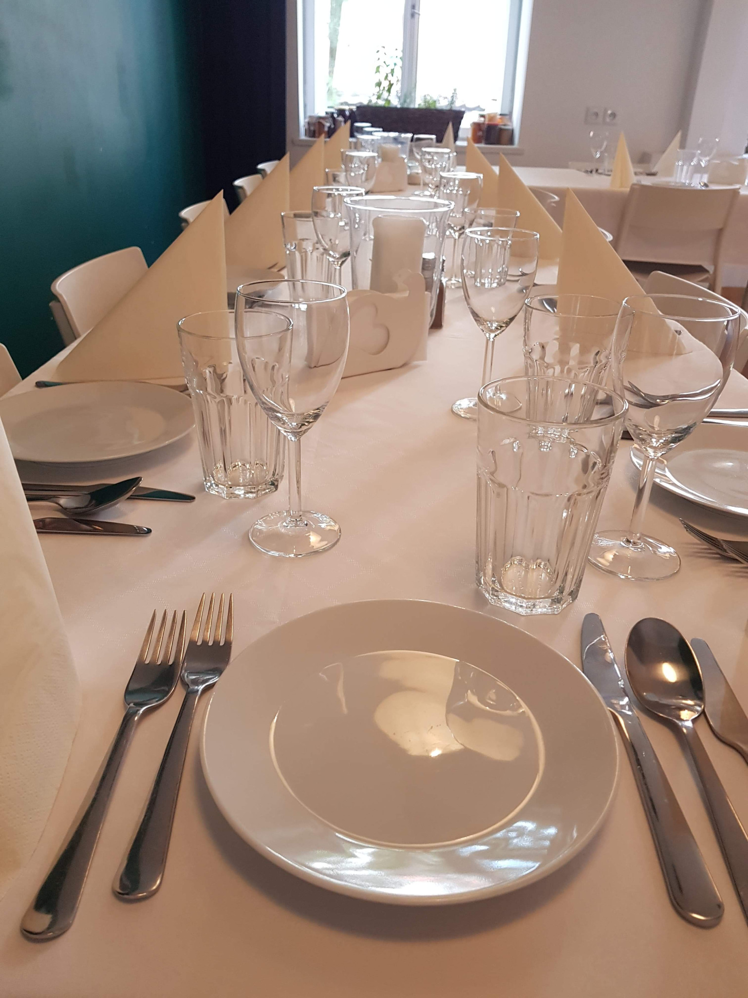 Elegancko nakryty stół z białym obrusem, talerzami, sztućcami, szklankami, kieliszkami i beżowymi serwetkami ułożonymi w trójkąty, przygotowany do przyjęcia gości.