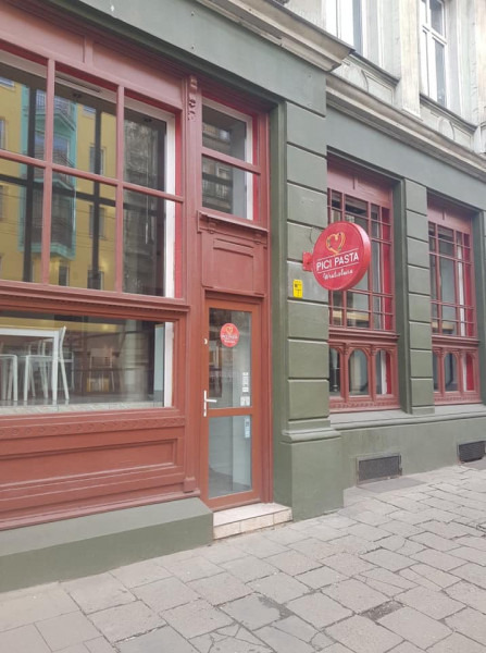 Fasada budynku z czerwonymi ramami okiennymi i szyldem 'Pici Pasta Tortellini' na zielonej ścianie, widok z chodnika.