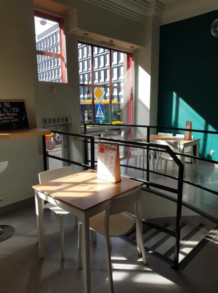 Jasne wnętrze kawiarni z widokiem na ulicę przez duże okna; stoliki z menu, czarne barierki oddzielające poziomy, oraz tablica z ofertą na ścianie.