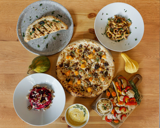 Kompozycja jesiennych dań na drewnianym stole: pizza z dynią i grzybami, calzone, pasta, sałatka z buraków i orzechów, dip dyniowy i warzywa na desce.