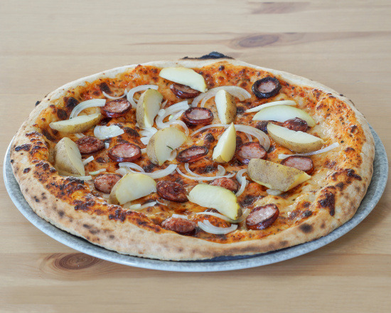Pizza z kiełbasą, ziemniakami i cebulą, ułożona na szarym talerzu na drewnianym stole, z widocznymi przypalonymi brzegami ciasta.