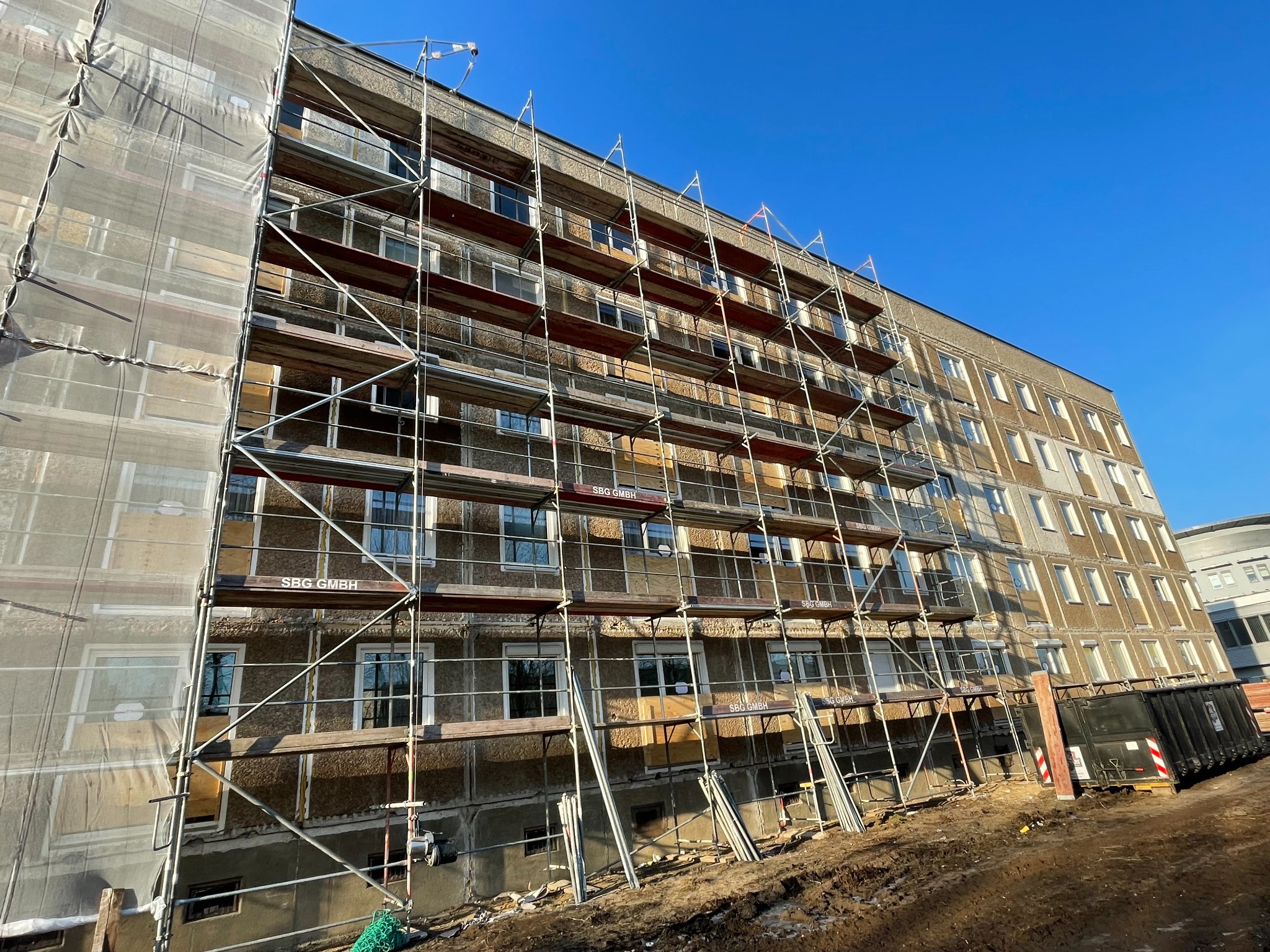 Wielopiętrowy budynek w trakcie renowacji, osłonięty rusztowaniem, częściowo przykrytym siatką ochronną, widoczny kontener na gruz.